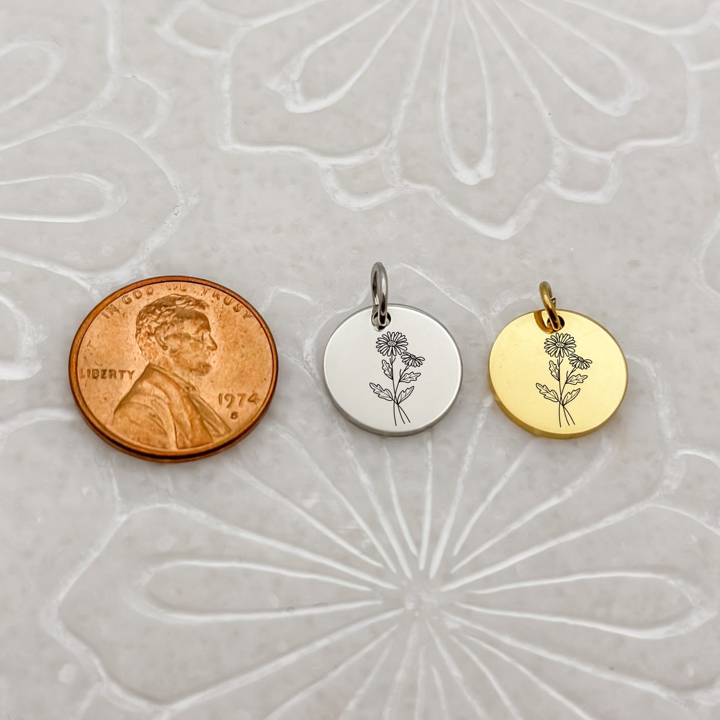 A detailed close-up of two April birth flower charms—one in 18K PVD Gold and one in Silver—engraved with a delicate daisy. Placed next to a penny for size reference.