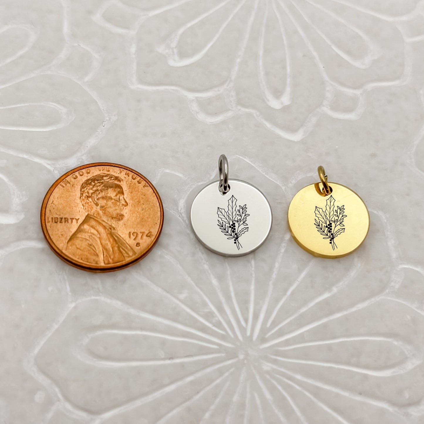 A detailed close-up of two December birth flower charms—one in 18K PVD Gold and one in Silver—engraved with a delicate holly branch. Placed next to a penny for size reference.
