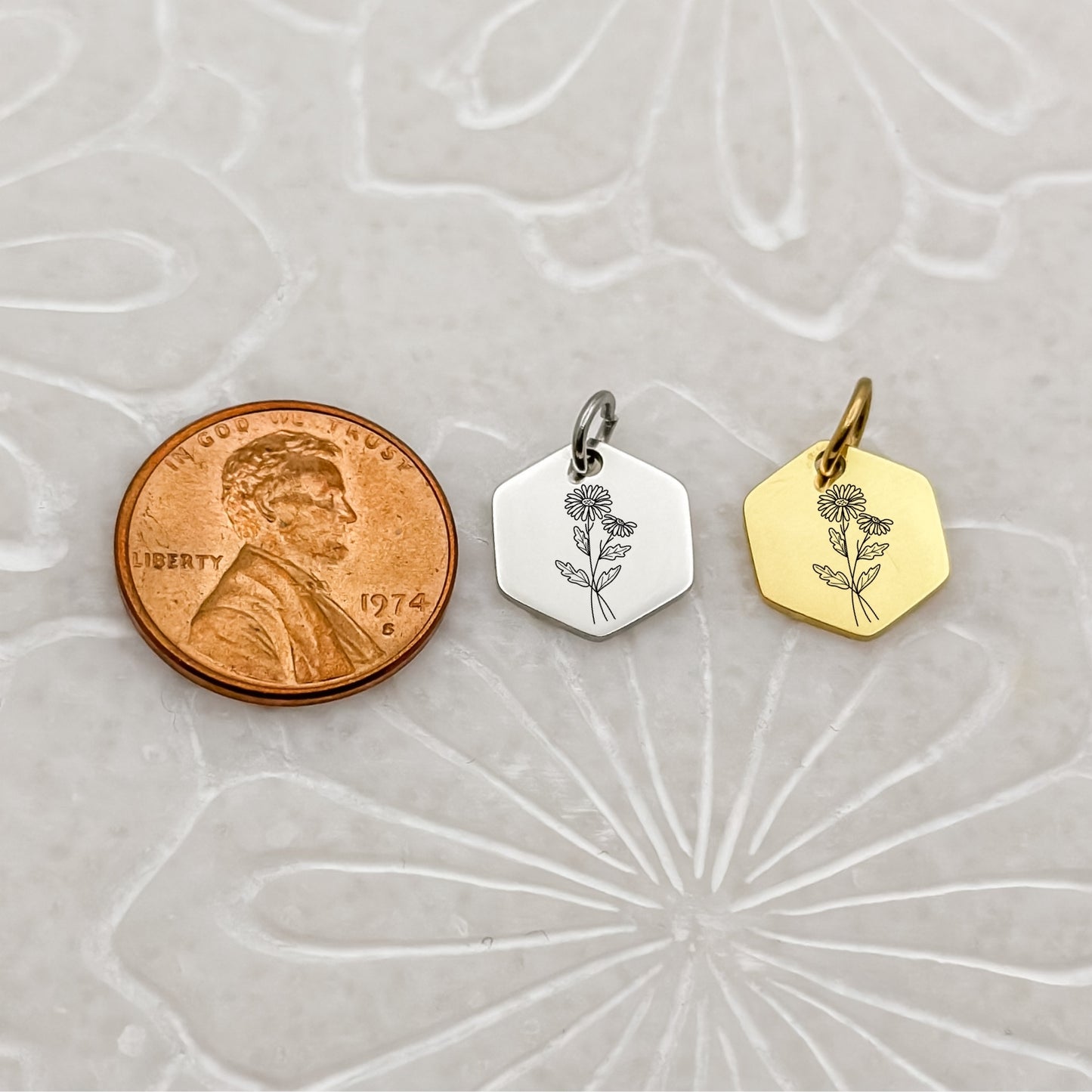 A close-up of two April birth flower hexagon charms in 18K PVD Gold and Silver, engraved with a minimalist daisy. Positioned beside a penny to show size.
