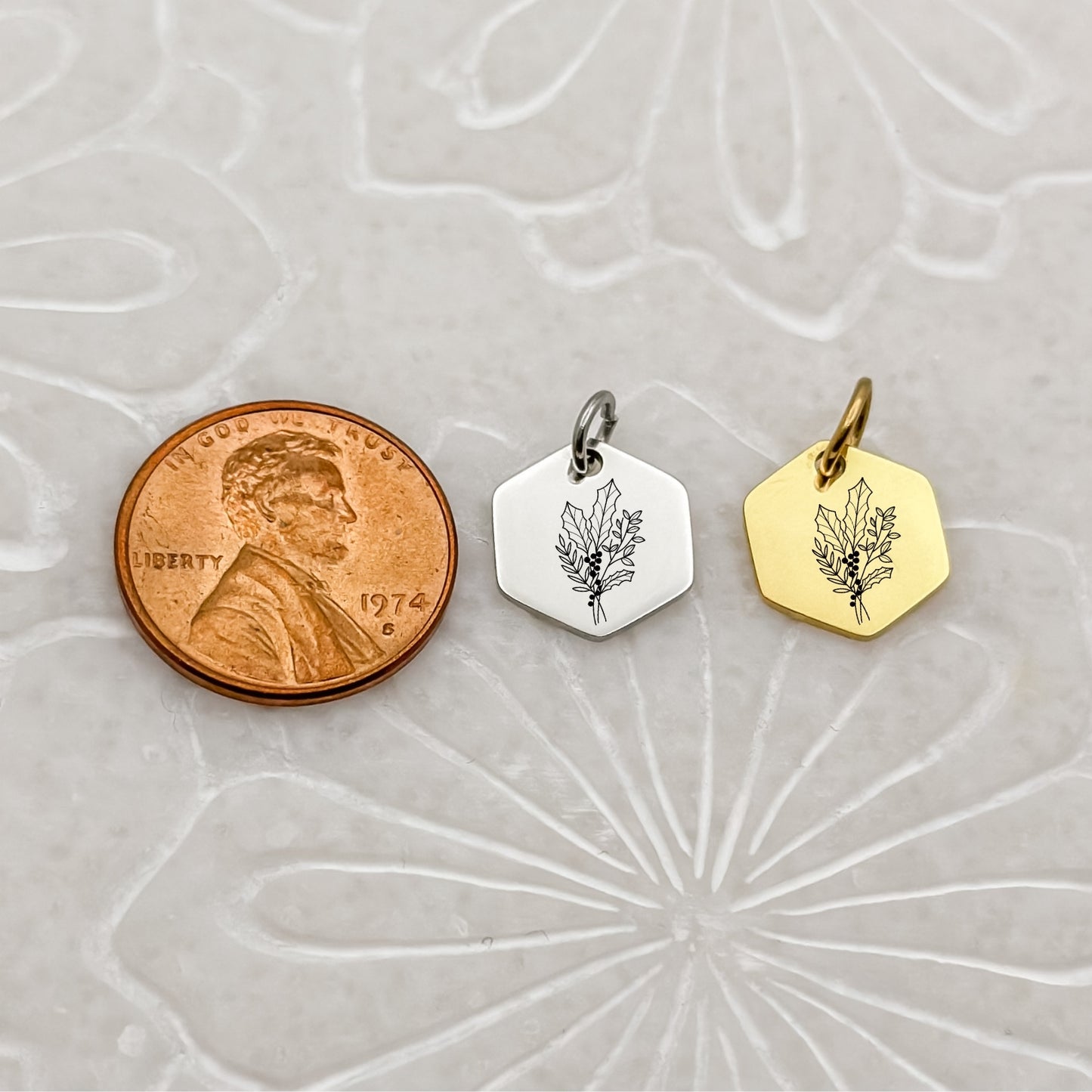 A close-up of two December birth flower charms in 18K PVD Gold and Silver, engraved with a minimalist holly branch. Positioned beside a penny to show size.