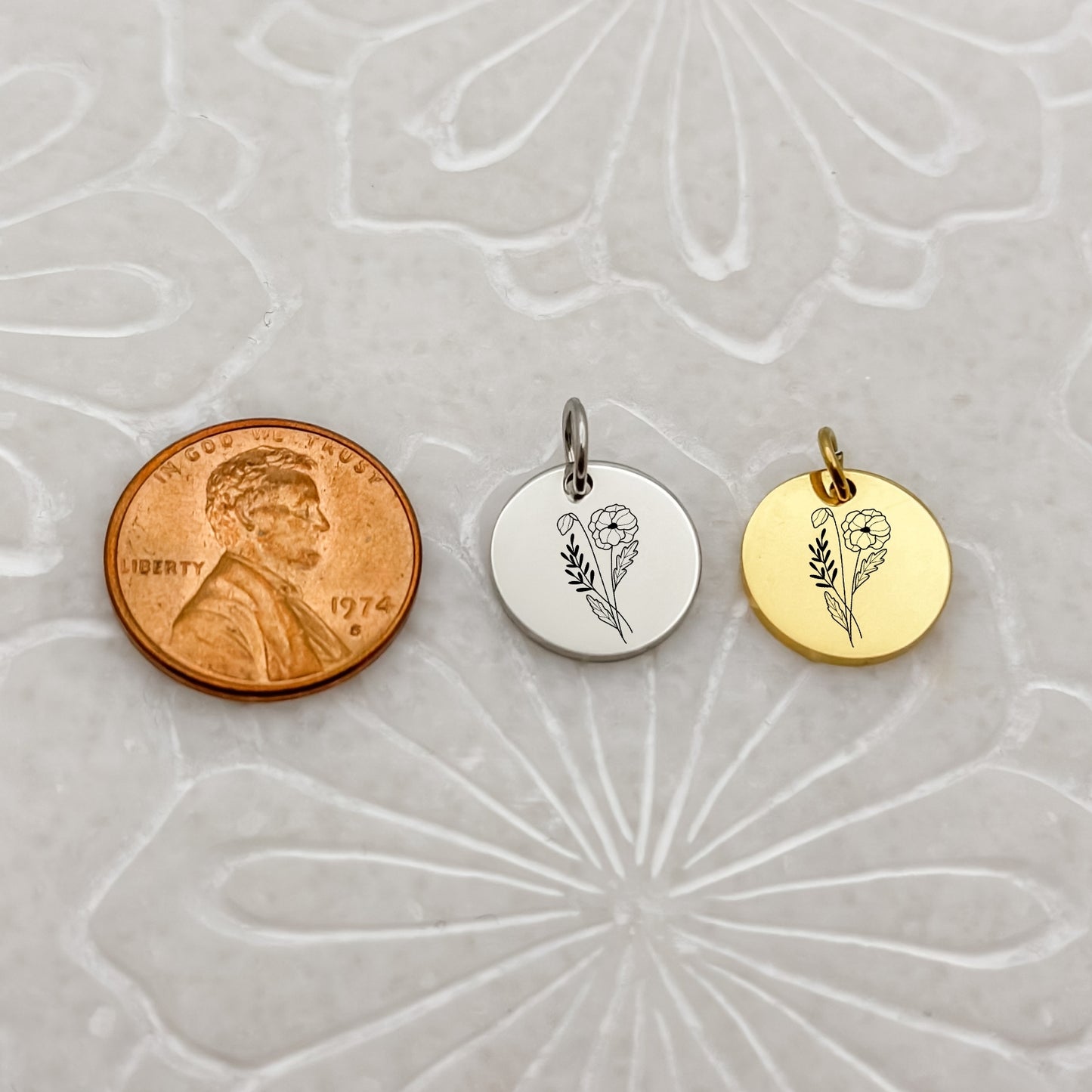 A detailed close-up of two August birth flower charms—one in 18K PVD Gold and one in Silver—engraved with a delicate poppy. Placed next to a penny for size reference.