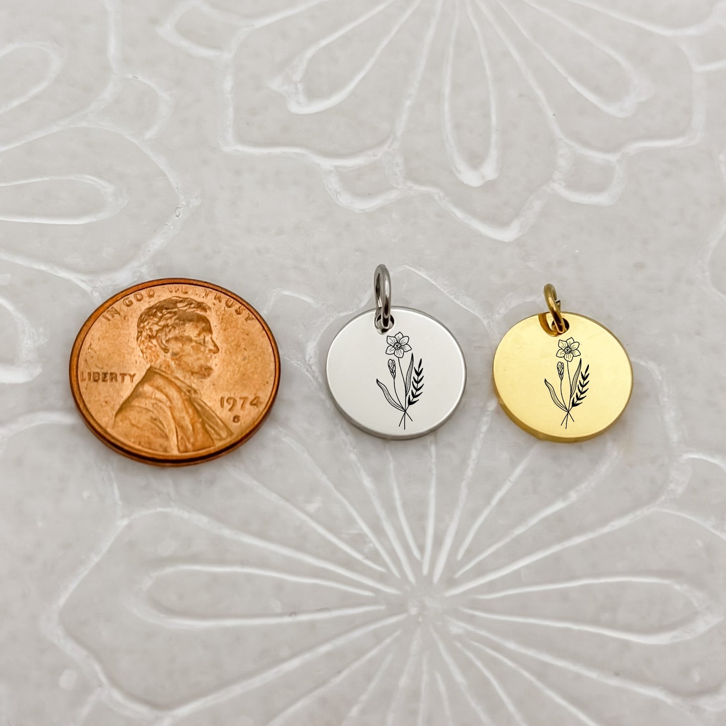 A detailed close-up of two March birthflower charms—one in 18K PVD Gold and one in Silver—engraved with a delicate daffodil. Placed next to a penny for size reference.