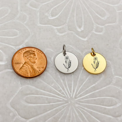 A detailed close-up of two March birthflower charms—one in 18K PVD Gold and one in Silver—engraved with a delicate daffodil. Placed next to a penny for size reference.
