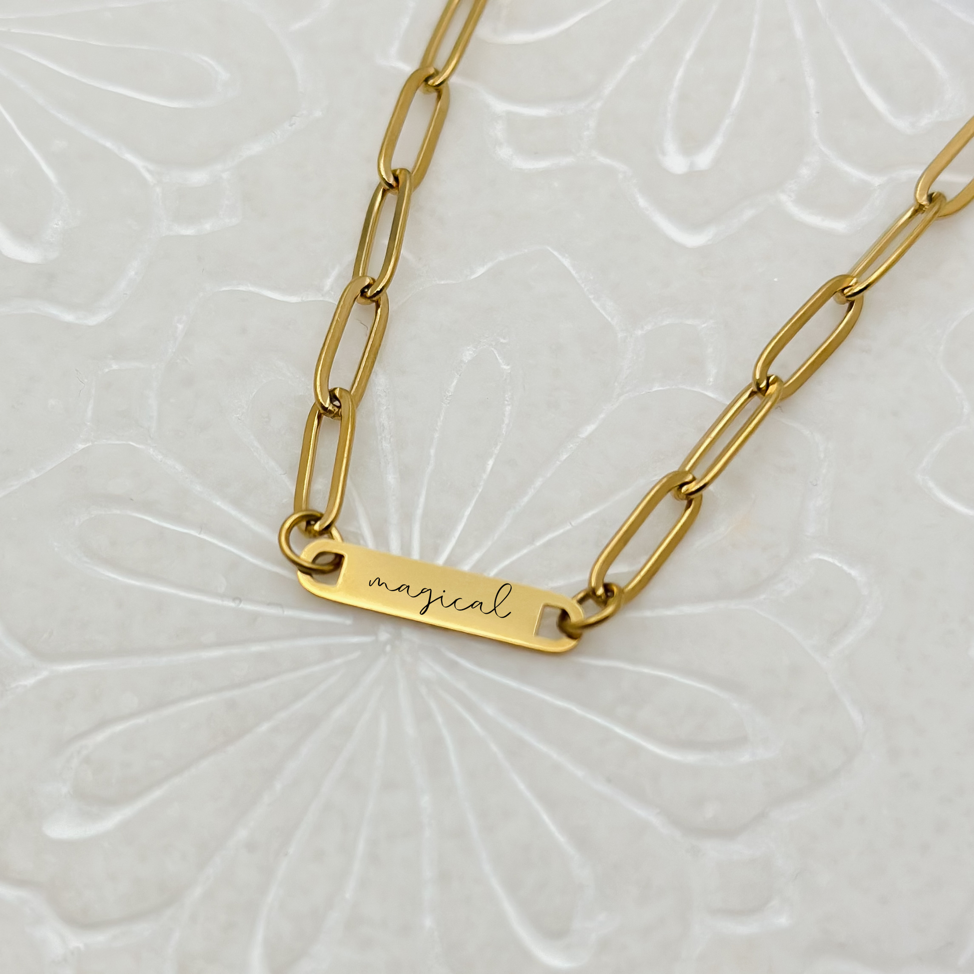 A minimalist gold bar necklace engraved with "magical" in script, displayed on a delicate paperclip chain.