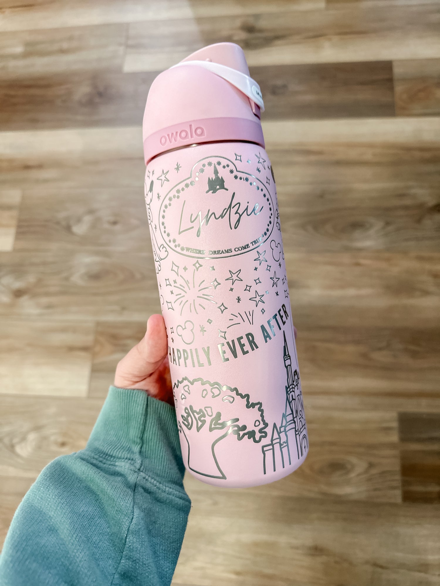 A hand holding a pink Owala water bottle with a whimsical "Happily Ever After" design. The bottle features magical illustrations, including fireworks, a castle, and a customizable name tag with "Lyndsie" written inside. The background shows a wooden floor