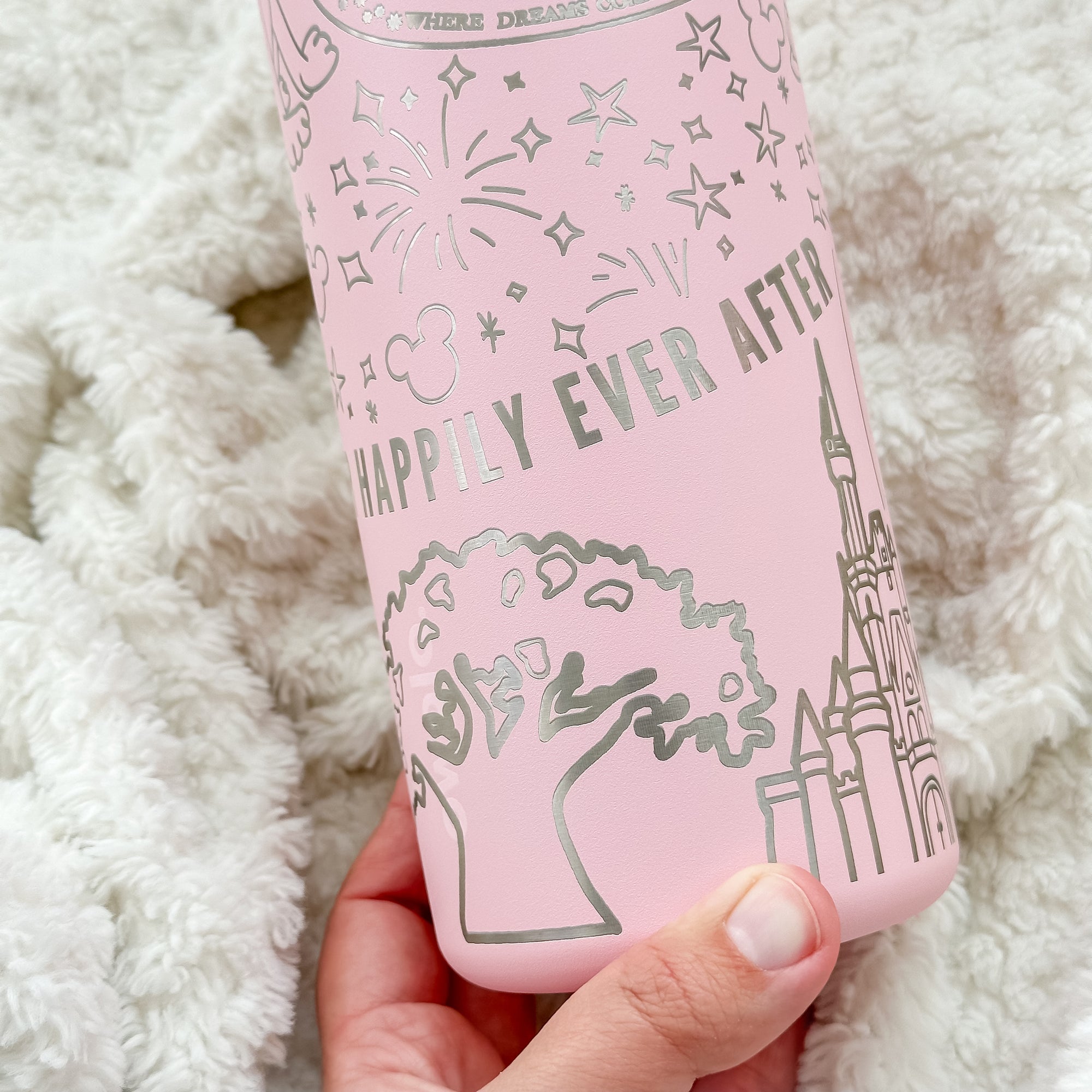 A close-up of a pink Owala water bottle featuring silver engraved details of fireworks, stars, a castle, a tree, and the phrase "Happily Ever After." The background is a soft, textured white blanket, and a hand is holding the bottle at the bottom.