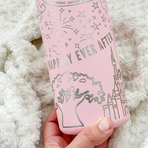 A close-up of a pink Owala water bottle featuring silver engraved details of fireworks, stars, a castle, a tree, and the phrase "Happily Ever After." The background is a soft, textured white blanket, and a hand is holding the bottle at the bottom.