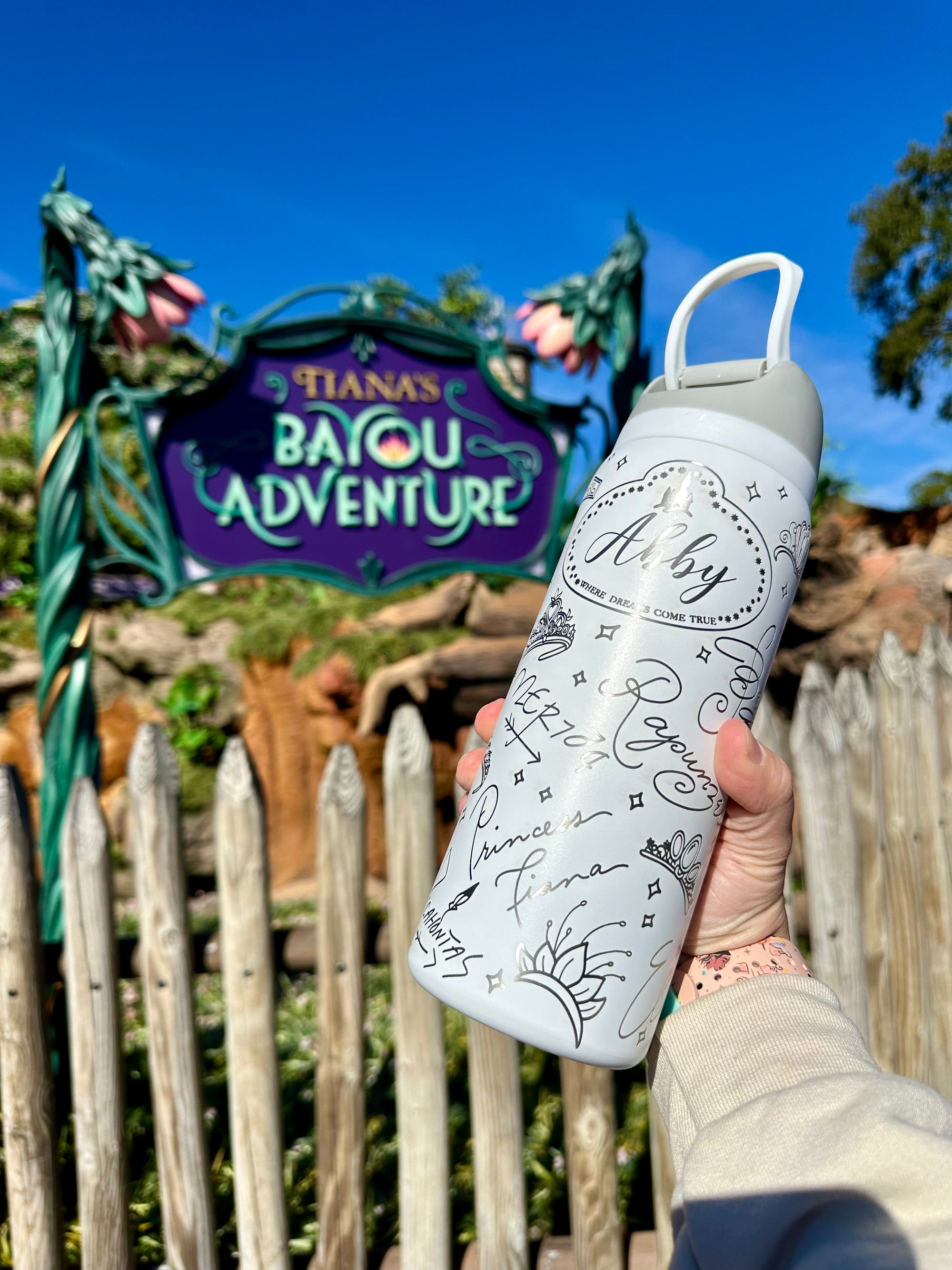 Person holding a water bottle with decorative text in front of Tiana's Bayou Adventure sign.