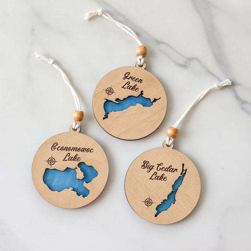 Three round wooden ornaments with lake illustrations and text on a marble surface
