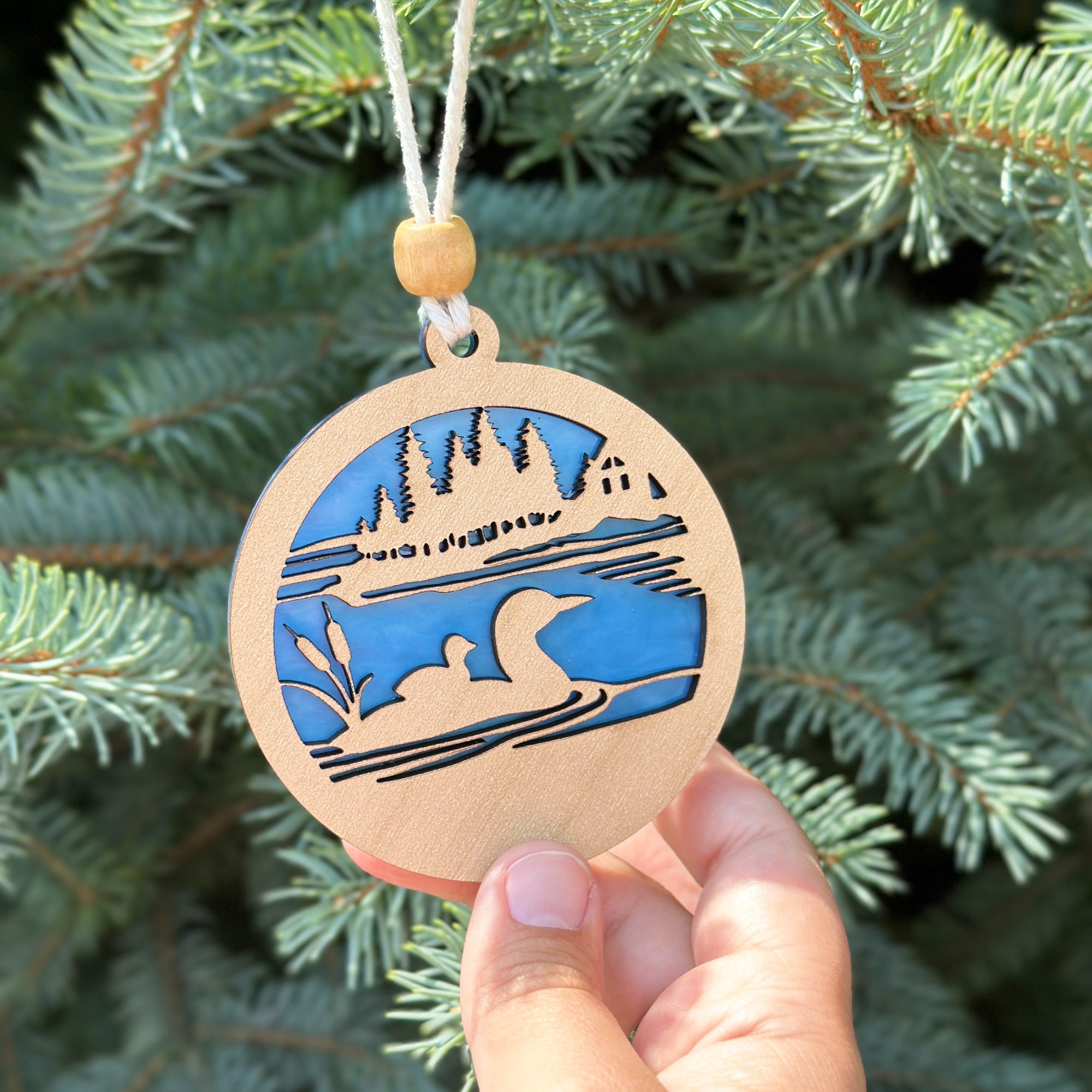 Wooden ornament with blue cutout design of a lake and mountains, held against a pine tree background.