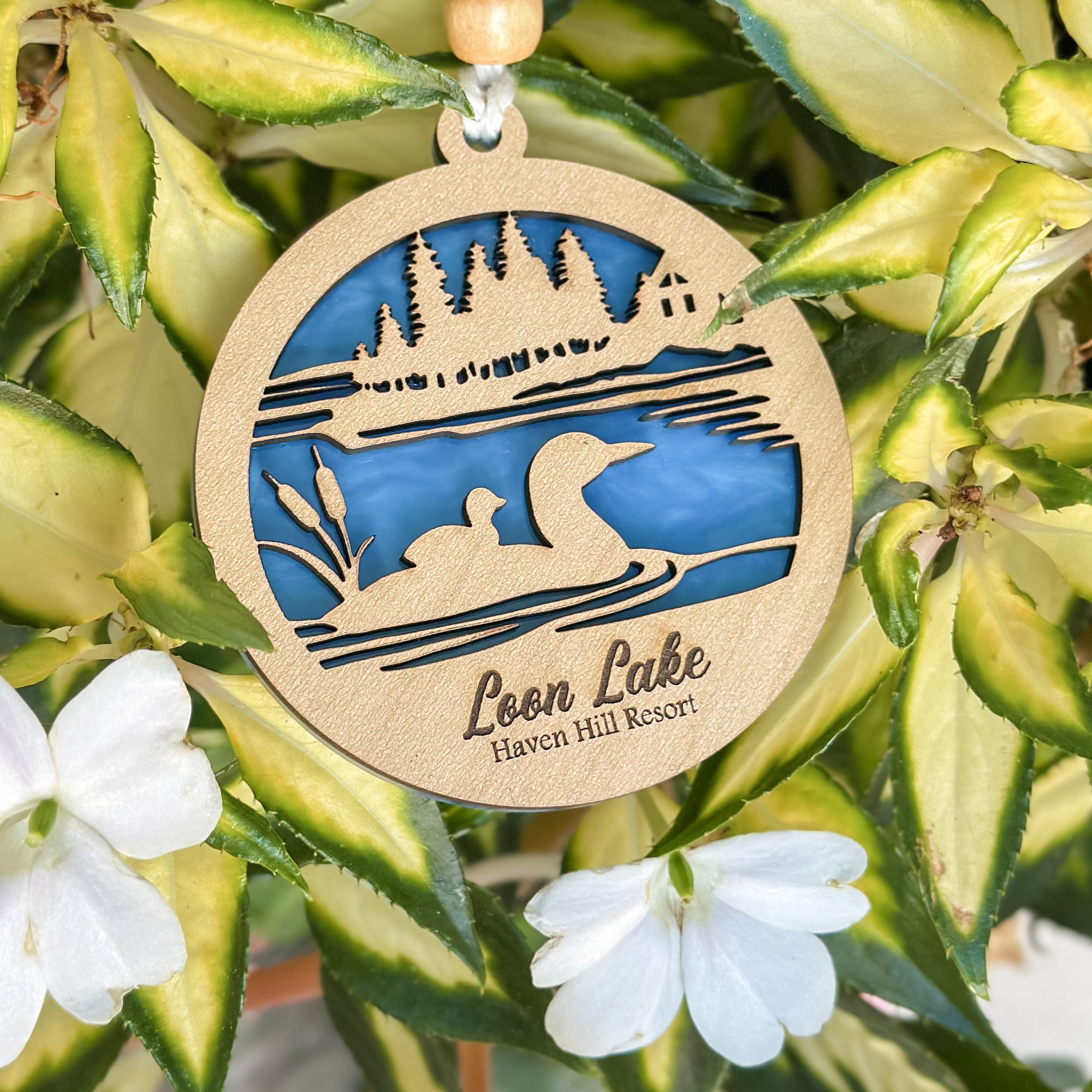 Decorative wooden ornament with lake scene and 'Loon Lake Haven Hill Resort' text, hanging among green leaves and white flowers.