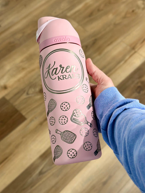 A pink 32oz Owala FreeSip water bottle with a custom engraved pickleball design and the name "Karen Kraus" in an elegant script inside a circular frame. The bottle features engraved pickleball paddles and balls, creating a stylish, personalized look. A person wearing a blue long-sleeve shirt is holding the bottle over a wooden floor background.