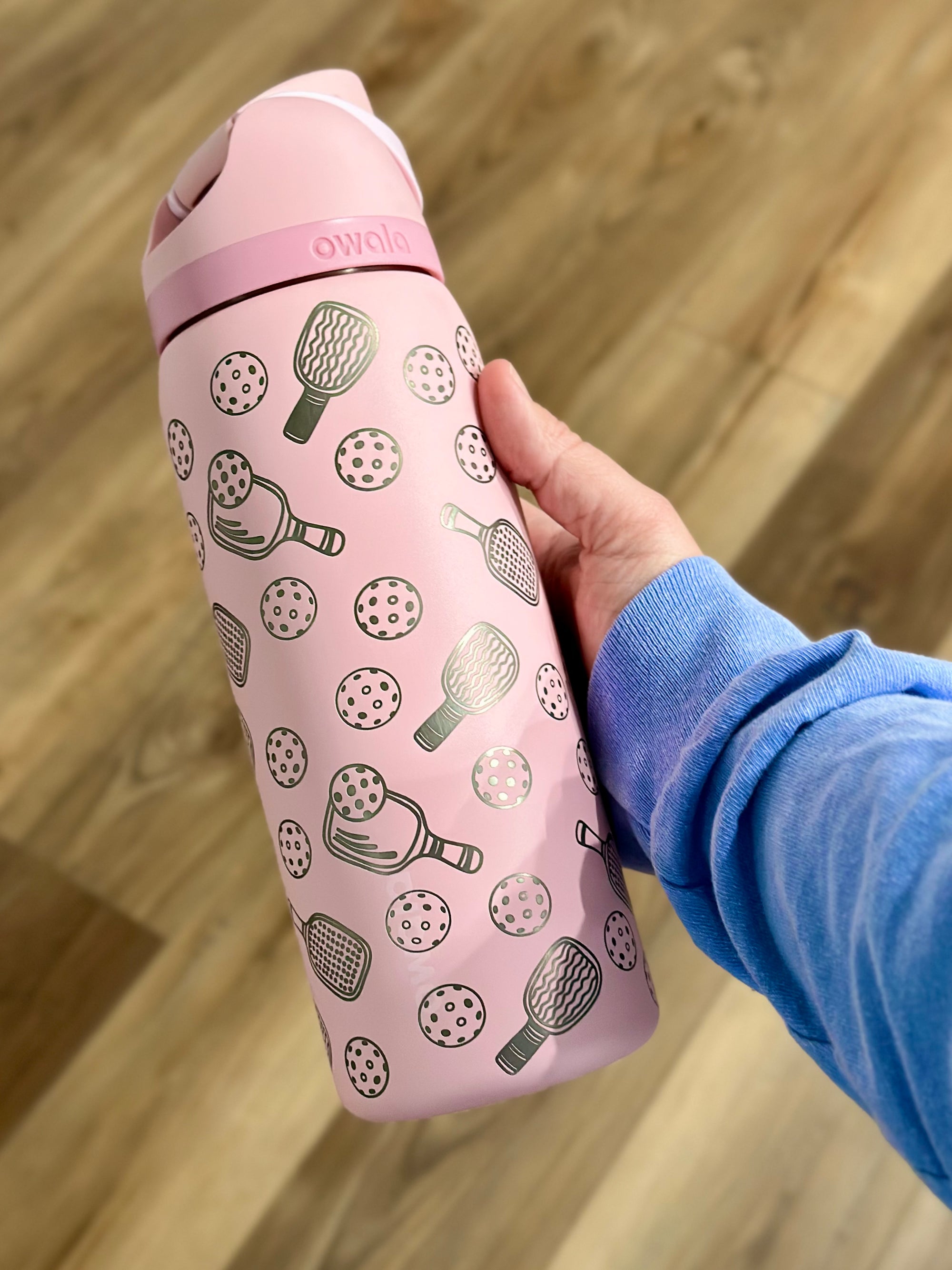 A pink 32oz Owala FreeSip water bottle with a custom engraved pickleball design and personalized name, displayed upright against a clean white background. The bottle features intricate engravings of pickleball paddles and balls, adding a stylish and sporty touch.
