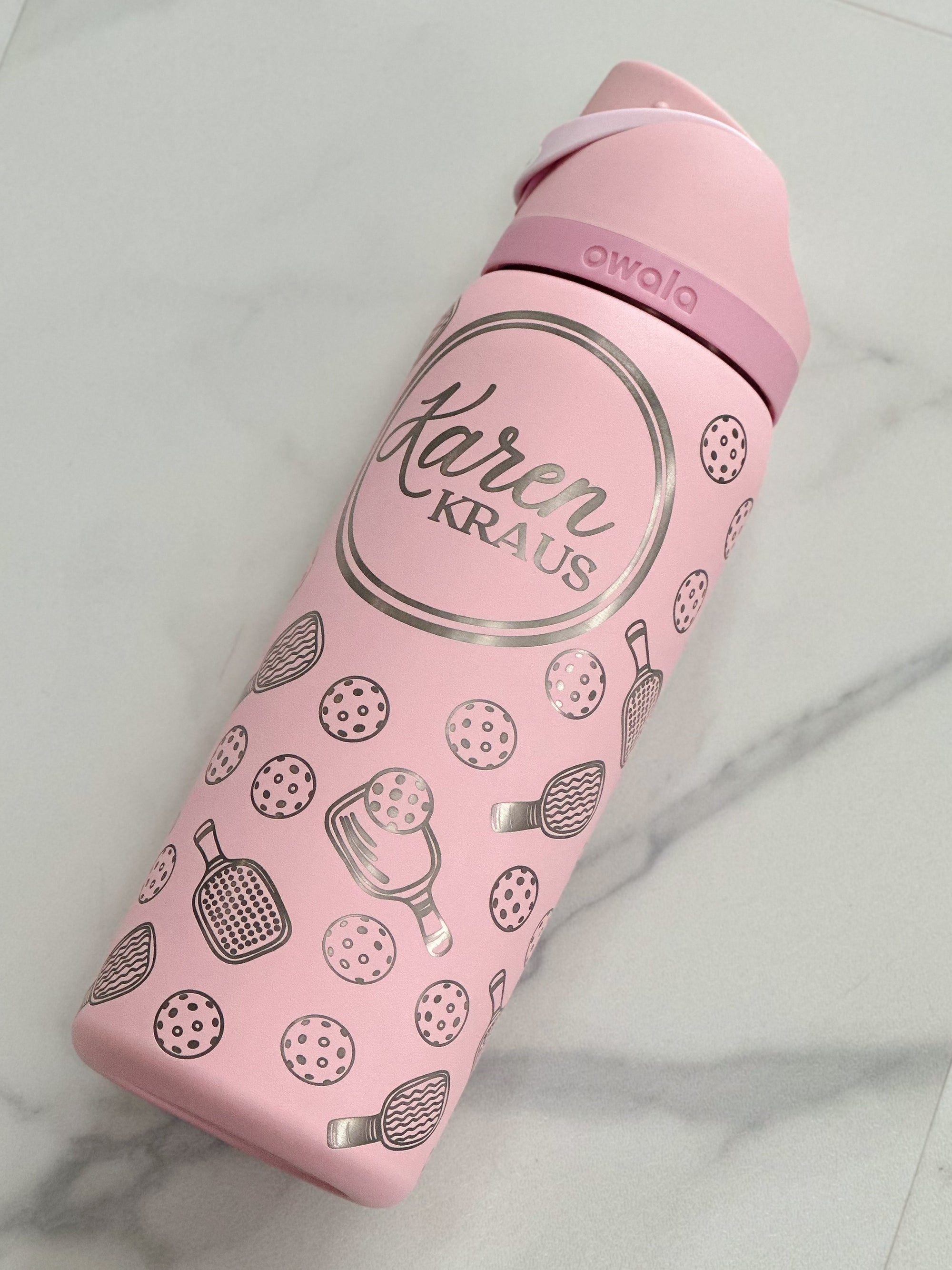 A 32oz pink Owala water bottle with a sleek pickleball-themed engraving, including paddles, balls, and a custom name inside a circular frame. The bottle stands against a marble background, highlighting its elegant and personalized design.