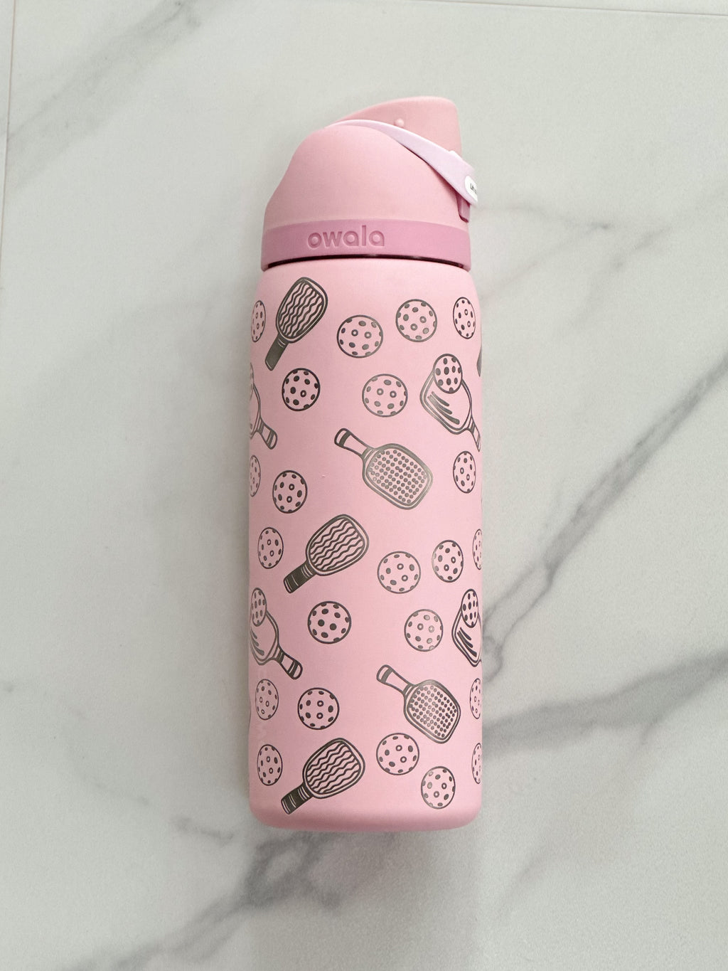 A personalized 32oz Owala FreeSip water bottle in soft pink, engraved with a pickleball motif and a custom name. Set against a crisp white backdrop, the design details stand out, making it a perfect gift for pickleball enthusiasts.