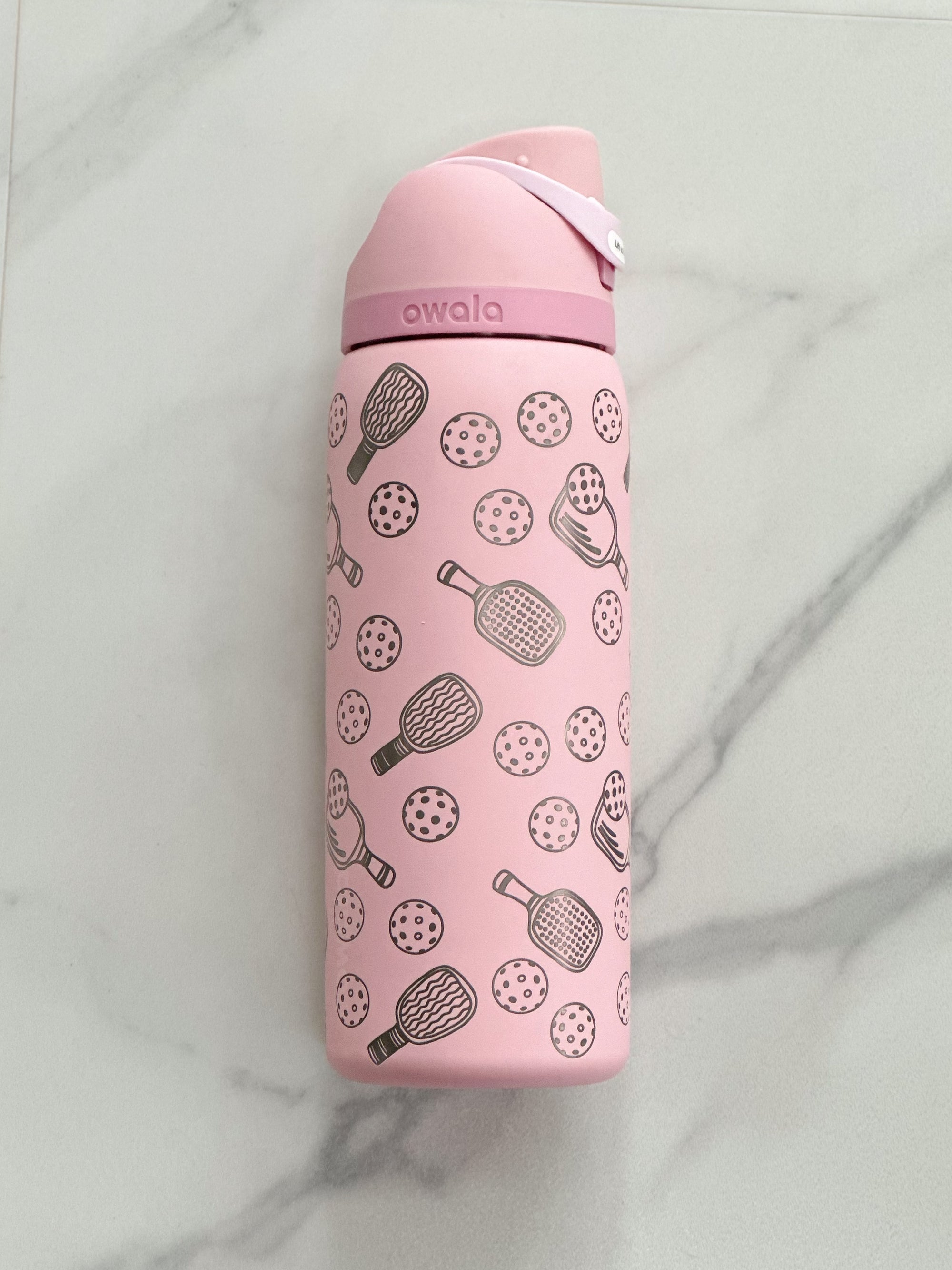 A personalized 32oz Owala FreeSip water bottle in soft pink, engraved with a pickleball motif and a custom name. Set against a crisp white backdrop, the design details stand out, making it a perfect gift for pickleball enthusiasts.