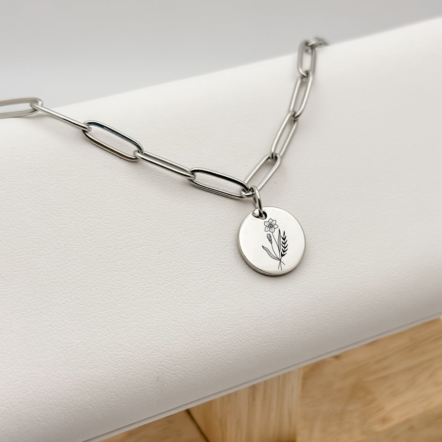 Silver circle charm necklace featuring a finely engraved daffodil, the March birthflower, on a modern paperclip chain. Hypoallergenic, waterproof, and tarnish-resistant.