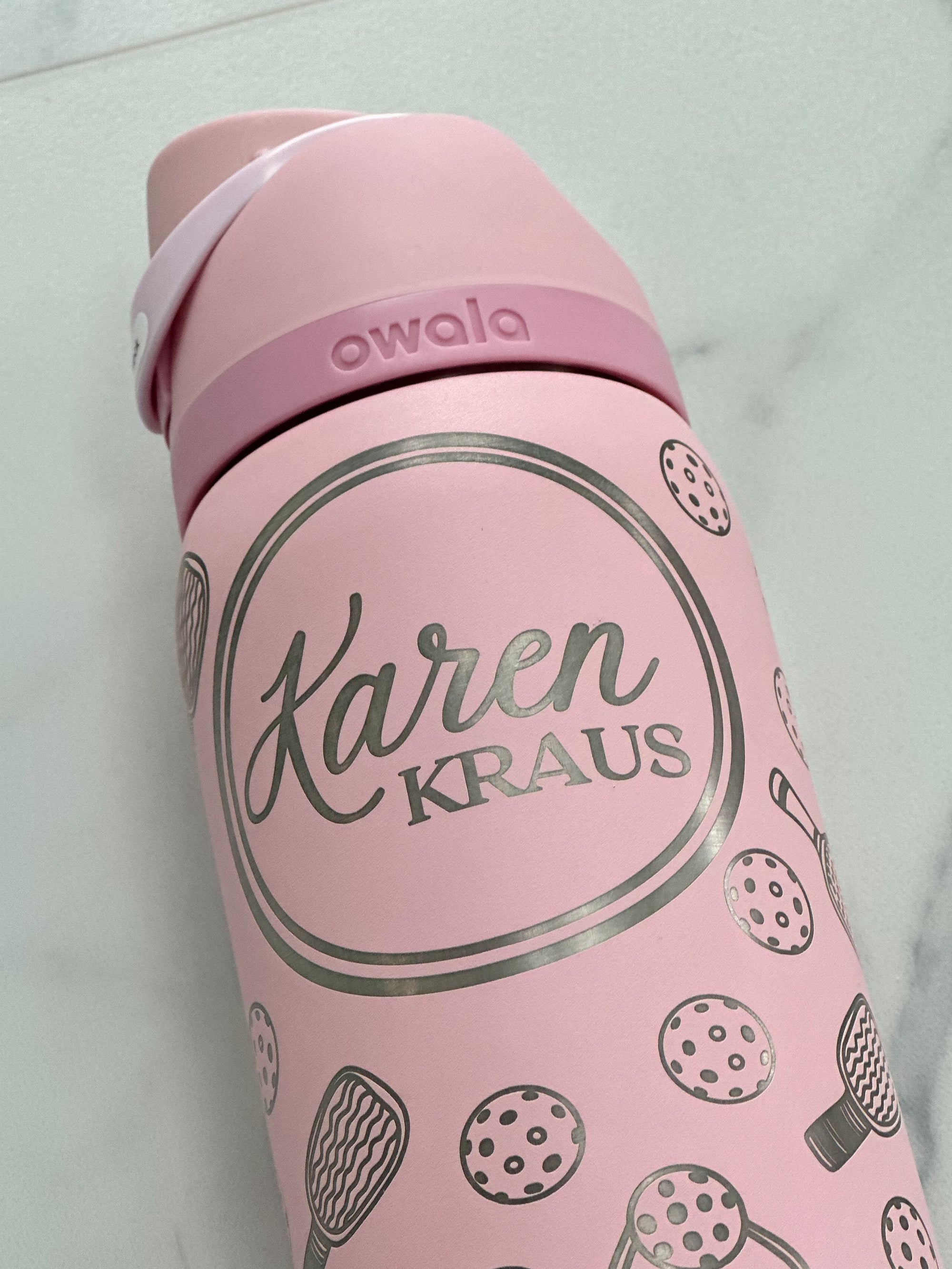 A personalized 32oz Owala FreeSip water bottle in soft pink, engraved with a pickleball motif and a custom name. Set against a crisp white backdrop, the design details stand out, making it a perfect gift for pickleball enthusiasts. Close Up photo