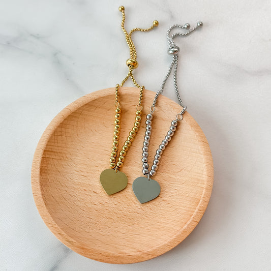 Two bracelets with heart-shaped pendants on a wooden dish against a marble background